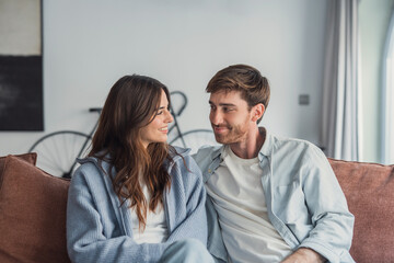 Obraz premium A young couple relaxes on a sofa, smiling and sharing a warm, intimate moment at home. Casual clothing, soft lighting and a bicycle in the background create a comfortable lifestyle scene