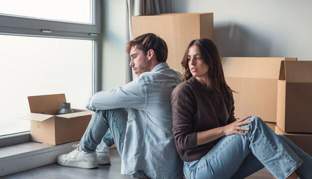 A young couple sits back-to-back among cardboard moving boxes by a window, appearing distant and tense after a move. The scene conveys stress, relationship strain, uncertainty and emotional fatigue
