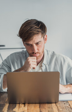 A focused young man concentrates on his laptop in a bright home office, working remotely and studying with thoughtful expression and a notebook nearby, conveying concentration, productivity and modern