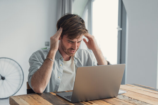 Young man experiencing frustration and headache while working on a laptop at a rustic wooden table in a bright home office, conveying stress, remote work challenges and digital fatigue