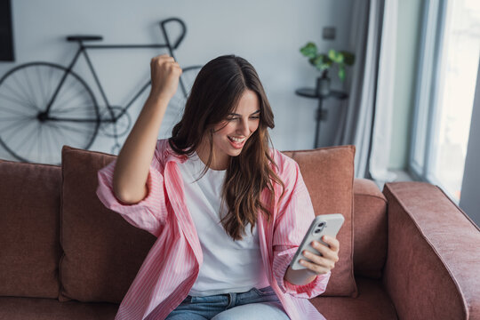 A young woman sits on a sofa in a bright living room, smiling and raising her fist while looking at her smartphone. Casual pink shirt, bicycle in background, celebrating a joyful online notification.