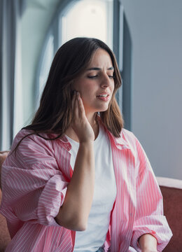 A young woman sits in a bright home interior, touching her jaw with a pained expression. The image suggests toothache, earache or TMJ discomfort and conveys worry, pain and health concern.