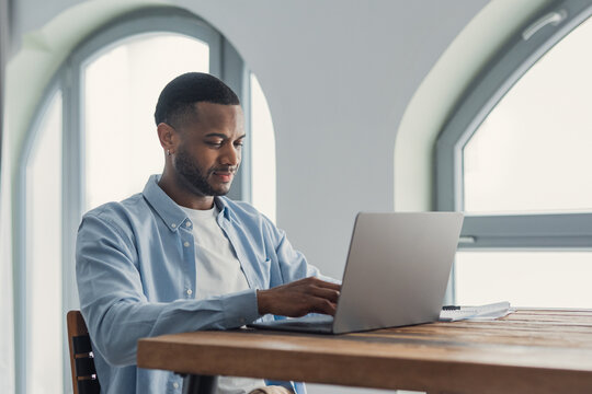A young man types on a laptop at a wooden desk in a bright home office, focused and calm while working remotely. Natural light, casual attire and a clean minimalist workspace convey productivity 