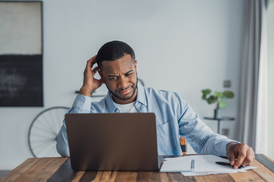 A young man sits at a wooden table in a home office, staring at his laptop with a puzzled expression while sorting paperwork and taking notes, conveying frustration and concentration during work