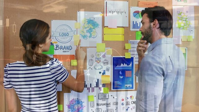 Man reaching sticky note on corkboard while woman arranging brand charts for marketing
