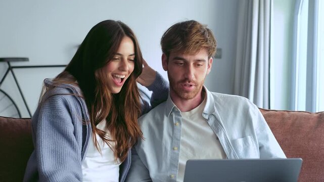 A happy young couple on a sofa at home shares an engaging moment while looking at a laptop. They smile, talk, and react with surprise and joy to what they see.