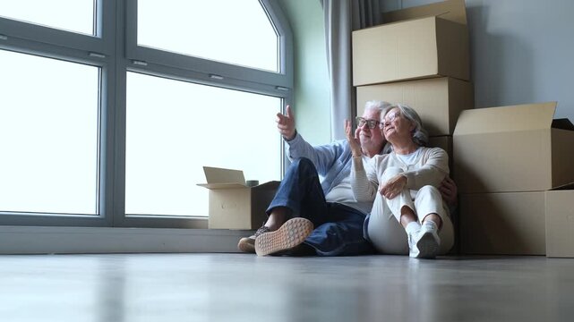 A happy senior couple sits on the floor of their new home, surrounded by moving boxes, as they discuss plans and envision their future together in this bright, empty space.