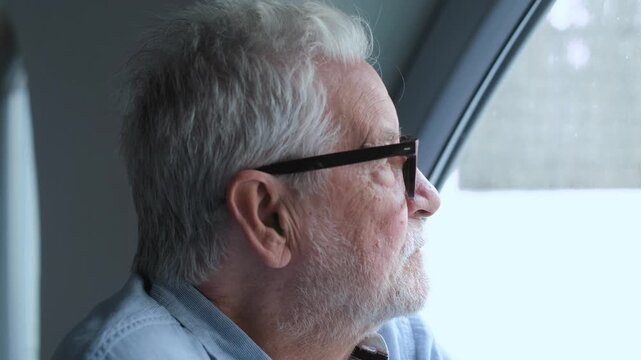 An elderly man with a beard and glasses gazes contemplatively out of a window. He reflects on life, perhaps thinking about memories or the future, showcasing a moment of quiet introspection.