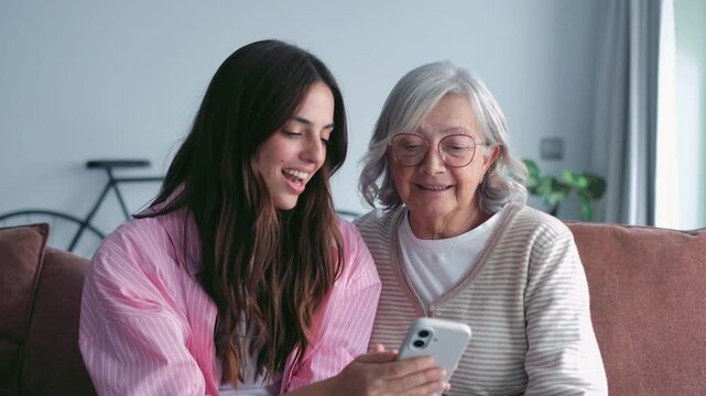 A young woman and her senior grandmother are sitting on a sofa, happily looking at a smartphone together. They are sharing a joyful intergenerational moment at home.