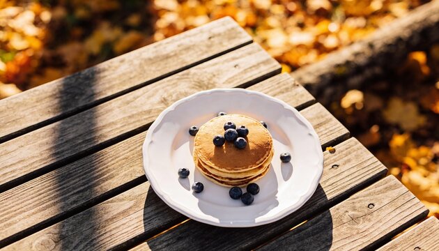 A stack of fluffy golden pancakes topped with fresh blueberries, served on a white plate atop a rustic wooden picnic table during a sunny autumn afternoon.