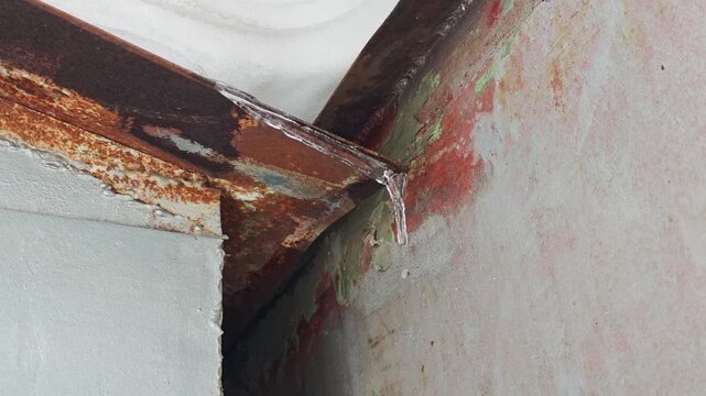 Water droplets flow from a rusted metal edge in a corner, revealing peeling paint and textured surfaces in an indoor setting with visible decay and wear