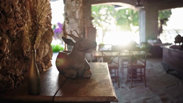A wooden deer sculpture displayed on a wooden table with morning sunlight streaming through. Coral stone wall background adds texture, creating a warm and artistic rustic atmosphere.