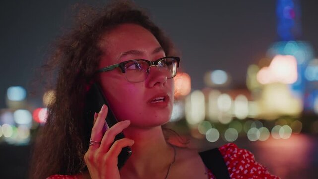 Young woman on phone by skyline, glasses framed face, neon bokeh lights, waterfront background, nighttime city atmosphere, contemplative expression, subtle glow, cinematic film grain, waiting