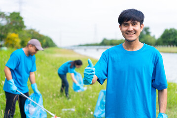Naklejka premium Volunteering, charity and clean environment concept. Male volunteers wearing blue t-shirt and protection glove standing and thumbs up to camera with volunteers diverse group cleaning park background.
