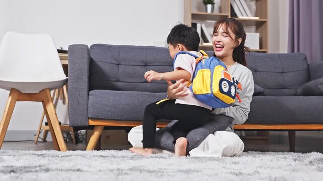 Asian mother young boy child son joyful female parent parenting living room grey sofa cozy home soft carpet family care comfort cheerful yellow backpack back to school playful hug relaxation bonding