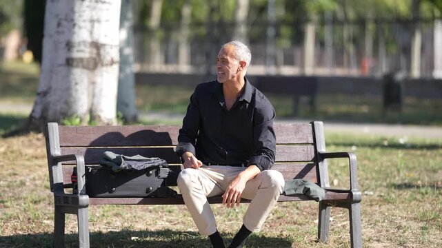 Man sits on park bench, relaxed, thoughtful. He gazes upward, eyes closed, relaxed posture. Sunlight filters through tall trees behind him. Green grass and trees surround the bench