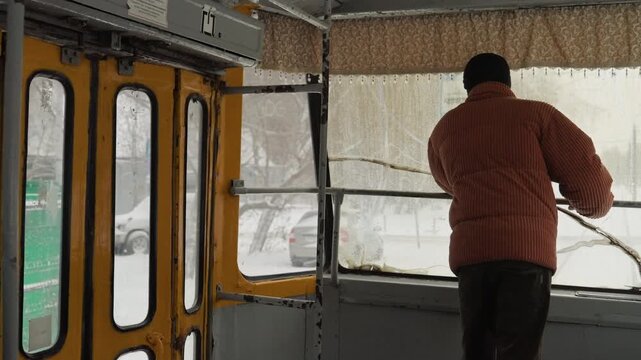 Man inspecting tram door amid snow, yellow interior panels frame scene, operator leans toward frosted glass and metal handle, parked cars and green fence visible outside, maintenance vibe with careful