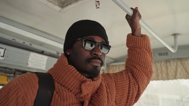 Black man gripping bus rail, wearing orange chunky sweater and sunglasses, scanning frosted cityscape through window backpack hints at creative professional headed to studio, assertive posture,