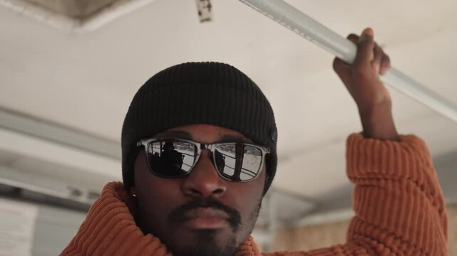 Black man knit beanie sunglasses, profile portrait inside bus holding overhead rail, reflective glass with cityscape, confident expression and layered orange sweater, backpack visible, soft diffused