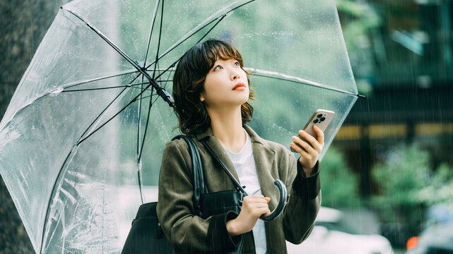 雨の日に傘を差しながらスマートフォンを使う日本人女性・ビジネスウーマンのビジネスイメージ, Japanese Businesswoman Using Smartphone While Holding an Umbrella on a Rainy Day