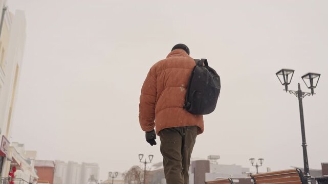 Black man walking away on snowy street wearing rust parka black backpack beanie and mittens walking past lampposts cityscape under overcast sky footsteps on frostcovered pavement panoramic back view
