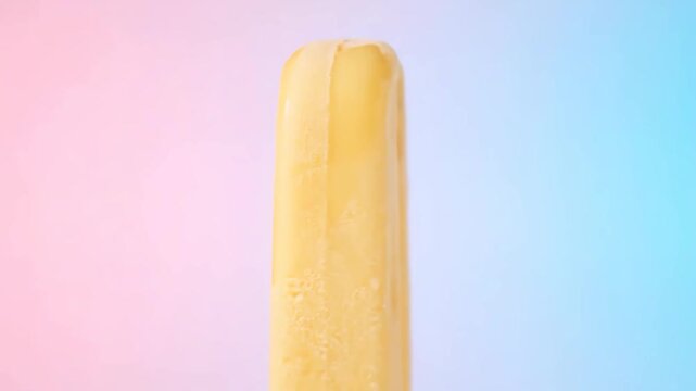 Close-up of vibrant yellow popsicle melting and dripping against a stylish pink and blue gradient background, a refreshing frozen summer dessert.