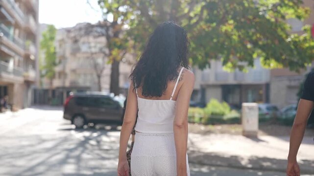 Sunlit street with three people walking together. Shadows drape the pavement beneath tall trees. Two women walk side by side, one in white, one in patterned dress