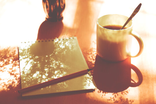 Shadow of vase with flowers, cup of coffee, notepad, pencil on the table with casting shadows. Inspiration and New story concept. Cozy inviting nook, quiet corner for inspiration