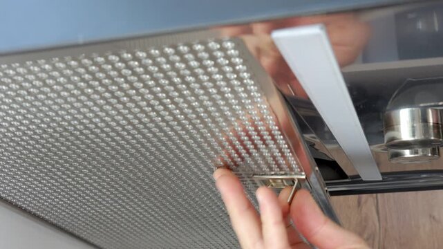 Hands removing metal filter from kitchen range hood in home interior. Concept of appliance cleaning household maintenance ventilation care and kitchen hygiene.