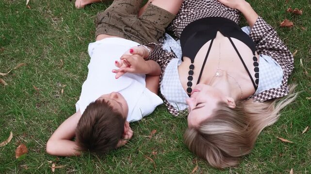 Family time. Outdoor leisure. Cheerful mother happy son in lush green grass holding hands relaxing enjoying summer weekend in park.