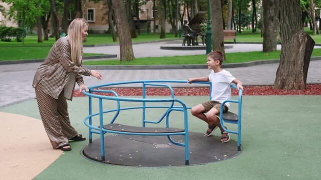 Family fun. Playground joy. Smiling positive mother happy son enjoying leisure together on blue merry go round in summer green park.