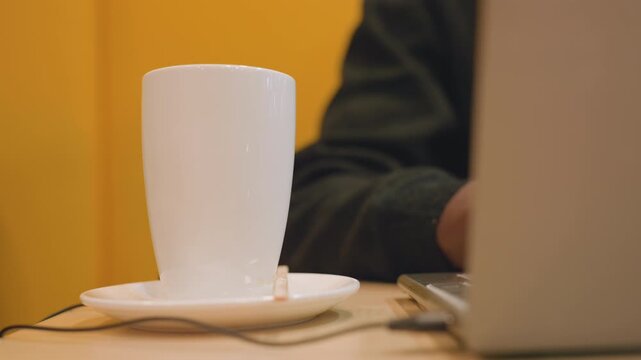 White cup beside black hands coding on laptop in vibrant yellow cafe, deadline pressure felt through keyboard clicks, USB cable and mouse visible, focused energy and caffeine boost, modern workspace