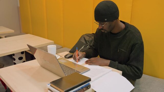 Black student studying at laptop desk, writing notes with pen, stack of textbooks and open notebook beside laptop, yellow acoustic panels, ceramic mug, quiet coworking study vibe, evening productivity