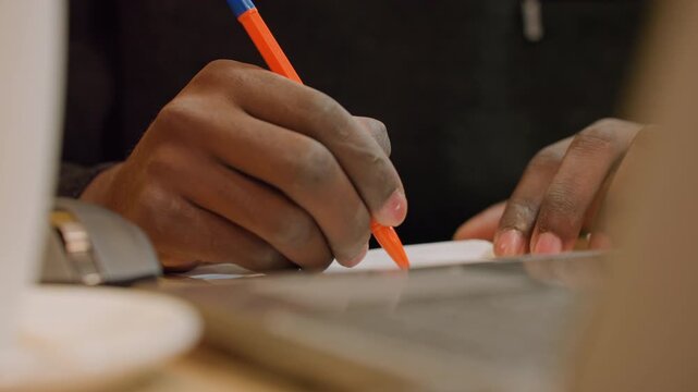 Closeup hands jotting notes by laptop in cafe remote worker capturing ideas with orange pen beside mug and saucer, ambient warm light, wristwatch visible, efficient morning hustle and planning.