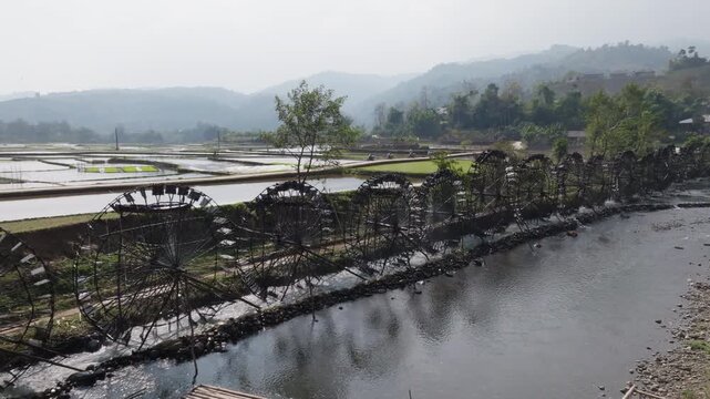 A line of bamboo water wheels stretches along the river at Guồng Nước N&agrave; Khương - Bản Bo, with rice paddies reflecting sunlight and mountains in the backdrop.