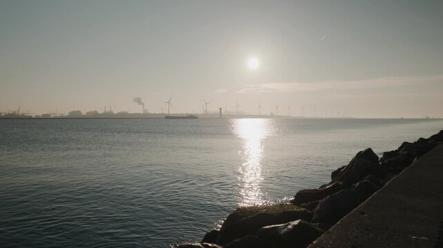 Dolly shot along the harbor entrance at Hoek van Holland with calm water sun reflection and distant wind turbines under clear sky