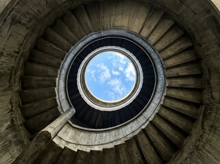 Fototapeta premium A view looking up from the bottom of a concrete spiral staircase toward a circular opening showing the bright blue sky with clouds
