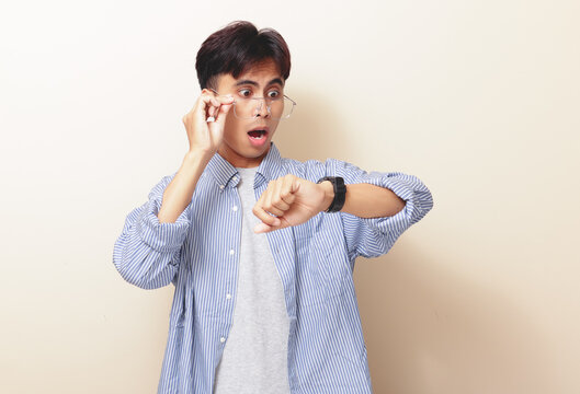 Funny shocked young Asian man looking at wristwatch with open mouth gesture on isolated background. Time management, late and deadline concept.
