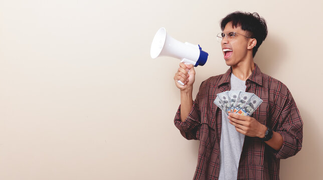 Excited young Asian man holding money banknotes and shouting into a megaphone on isolated background.