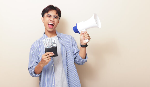 Very excited young Asian man holding wallet full of money and shouting into a megaphone on isolated background.