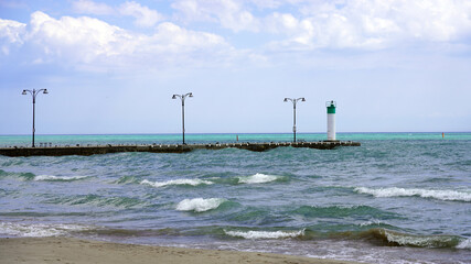  A small white lighthouse with a green top stands at the end of a stone pier extending into the rolling turquoise waves of Lake Ontario along the Oshawa lakefront © Gregory