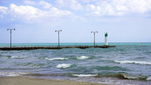 
A small white lighthouse with a green top stands at the end of a stone pier extending into the rolling turquoise waves of Lake Ontario along the Oshawa lakefront