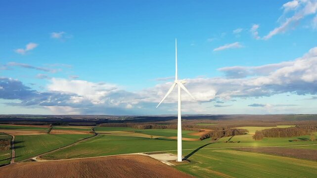 A single wind turbine stands in the middle of green agricultural fields under a clear blue sky in the Nivernais region near Oisy, France. The landscape features rolling farmland and distant forests