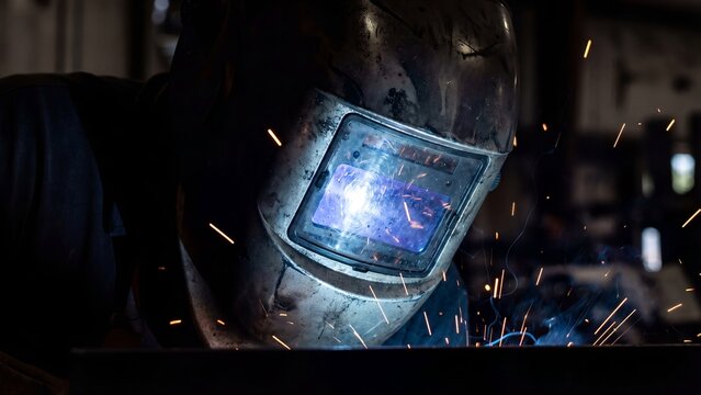A welder wearing a protective mask works diligently in a workshop with sparks flying everywhere closely focused on task