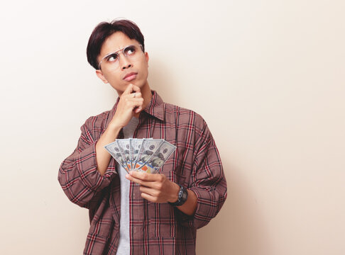 confused young asian man holding money touching his chin with thinking expression isolated background.