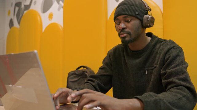 Black man typing on laptop focused in yellow coworking booth, headphones on, backpack at side, productivity and concentration during remote work session, casual modern setup with warm tones
