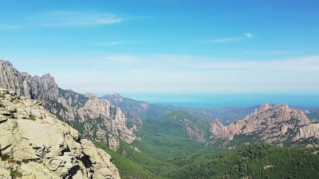 Scenic view of rugged mountains and lush forest at Col de Bavella in Corsica under a clear blue sky. The Mediterranean Sea is visible in the distance.