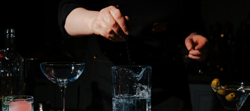 Woman bartender stirring martini ingredients in mixing glass with long bar spoon. Professional chilling and dilution technique for classic cocktail.