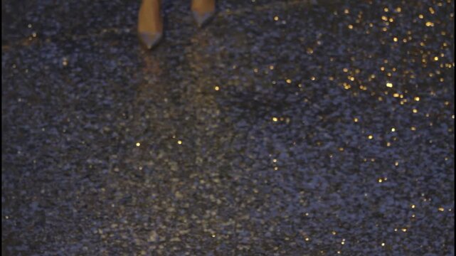 A woman's legs wearing metallic high heels and sequined pants on a glowing asphalt road at night.
