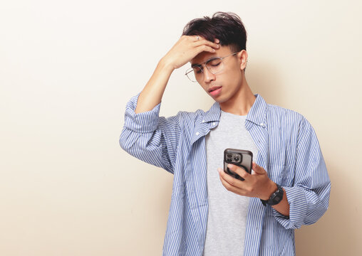 Stressed young Asian man holding smartphone and touching forehead with dizzy expression on isolated background.